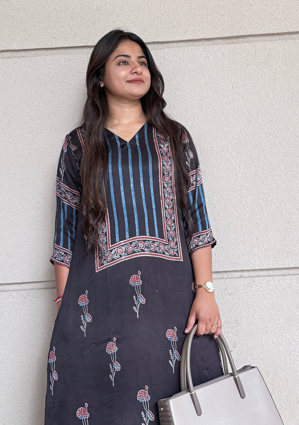 Woman in a dark floral dress holding a white handbag against a light gray wall.